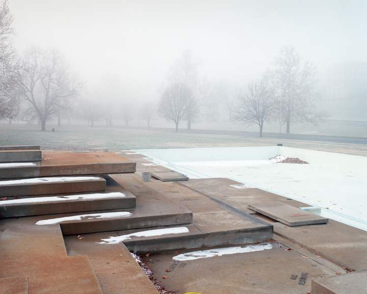 A misty park landscape featuring bare winter trees receding into fog, with a drained concrete wading pool in the foreground. Rusted metal structural elements and wooden beams lie across the pool's floor, creating geometric shadows on the pale surface.