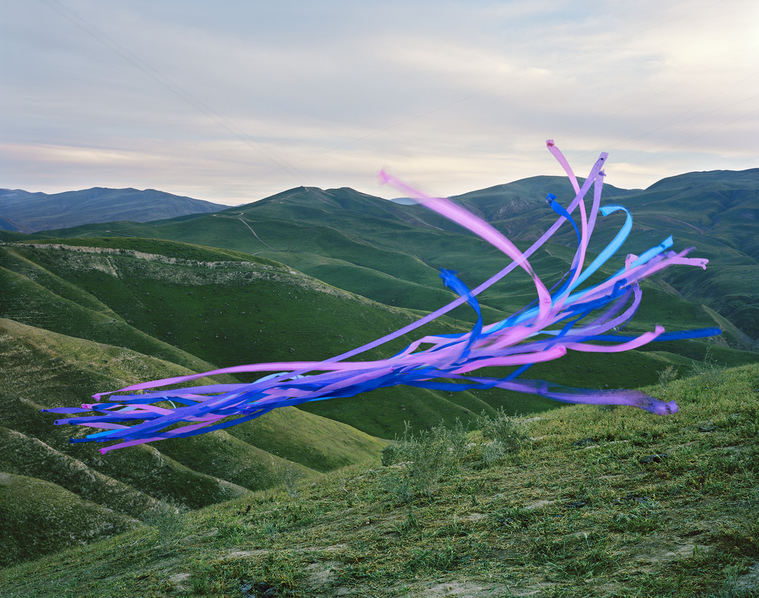 Vibrant neon-colored streamers in shades of blue, pink, and purple cascade across a rolling landscape of green hills under an overcast sky, creating a striking contrast between the digital-looking synthetic forms and the natural terrain.