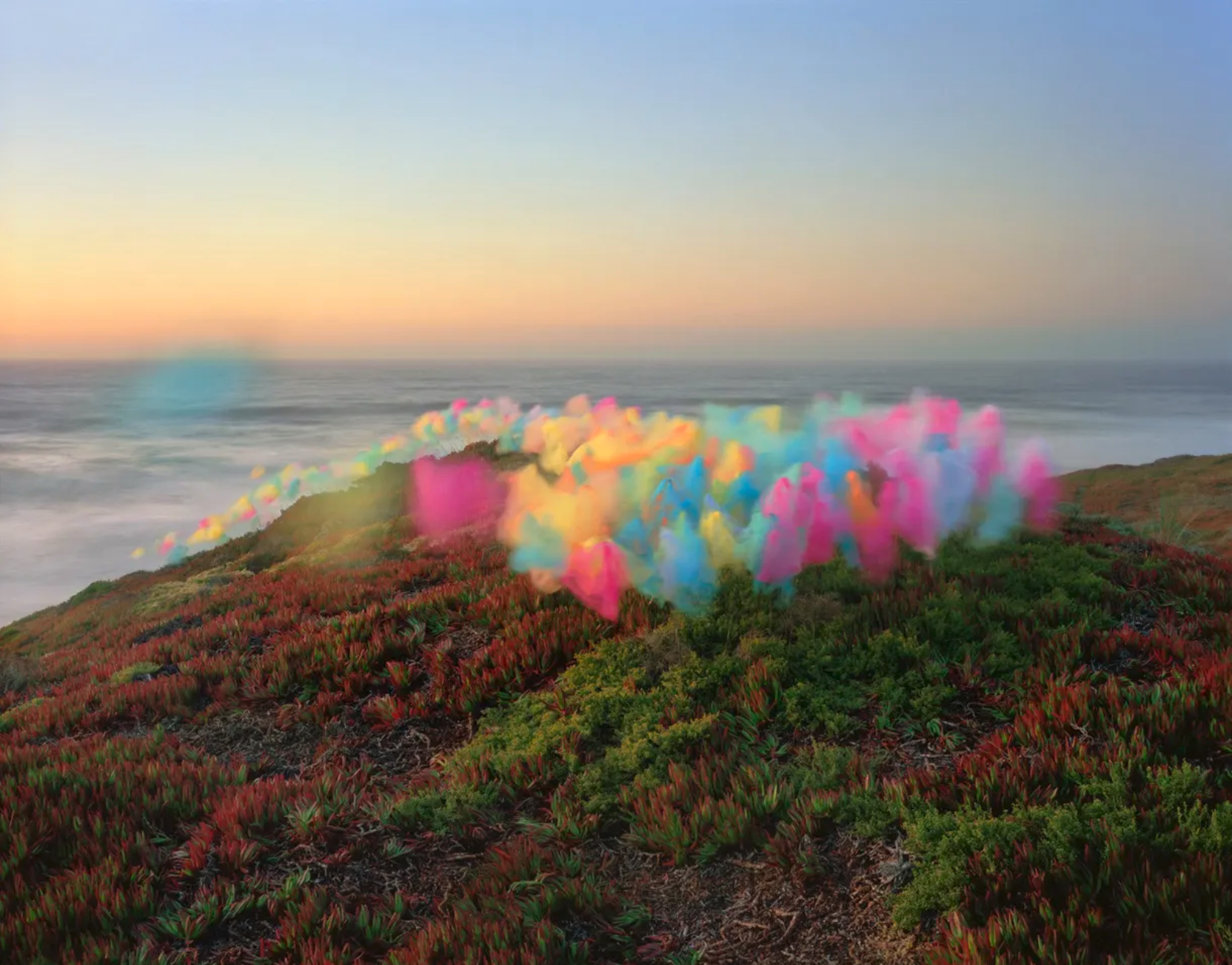 Coastal hillside densely covered in red and green vegetation overlooking a misty ocean at dawn, with a billowing cloud of vibrant rainbow-colored tulle fabric suspended across the landscape.