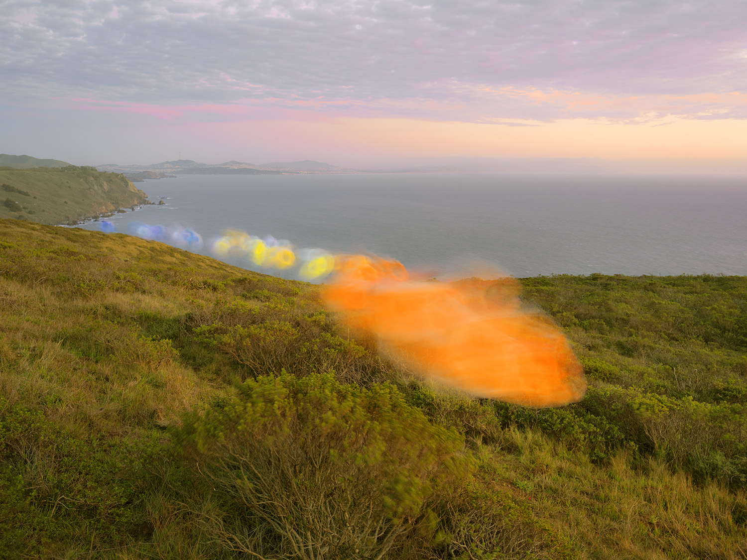 Coastal landscape with green hillside vegetation in foreground, expansive ocean and distant headlands under soft twilight sky, with an orange and yellow tulle fabric stretched across the middle ground creating an ethereal, translucent veil over the seascape.