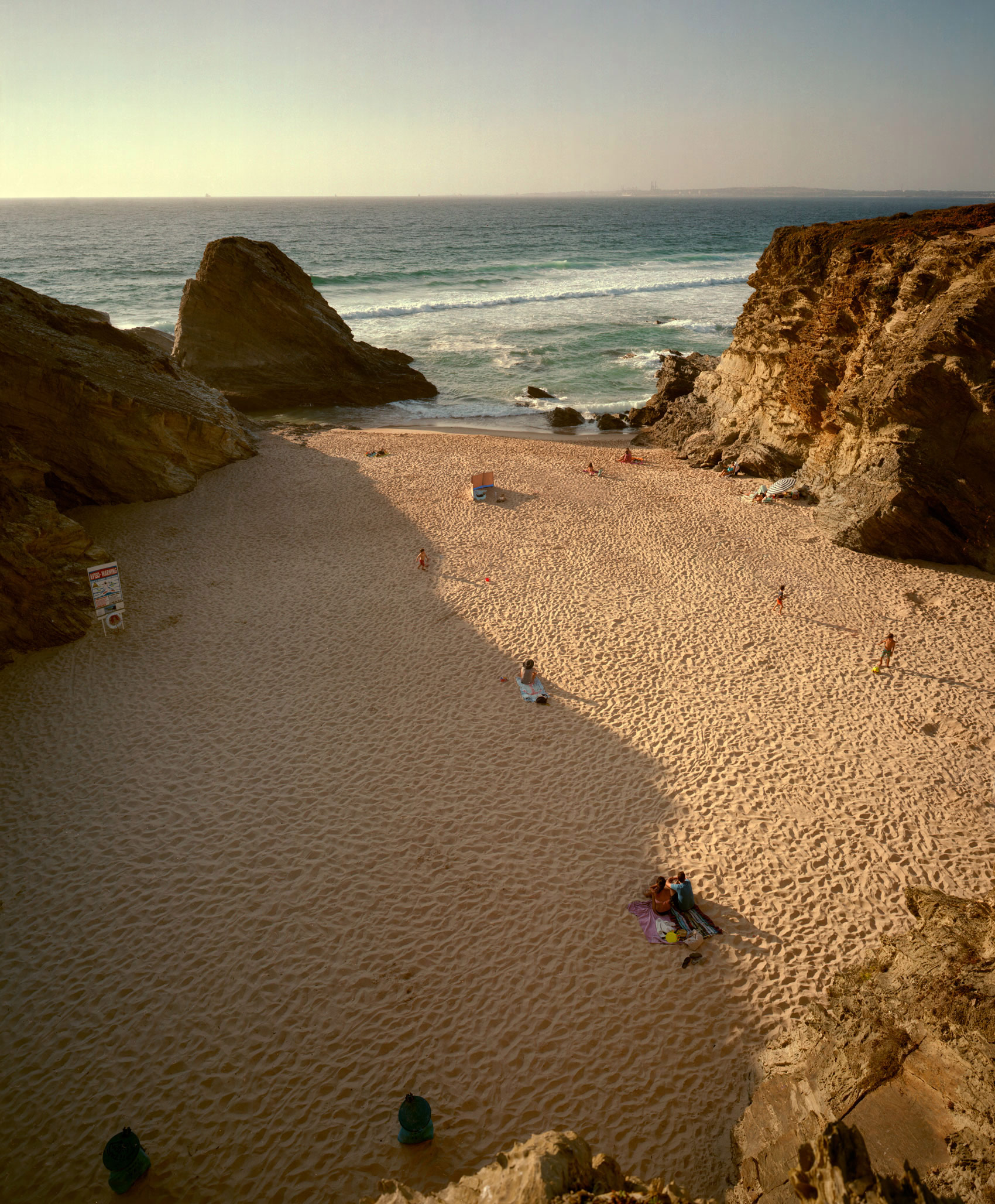 A secluded beach cove framed by towering ochre-colored cliffs, with golden sand and turquoise waters. Several beachgoers are scattered across the expansive shoreline, their small figures emphasizing the dramatic scale of the rocky formations and vast seascape beyond.