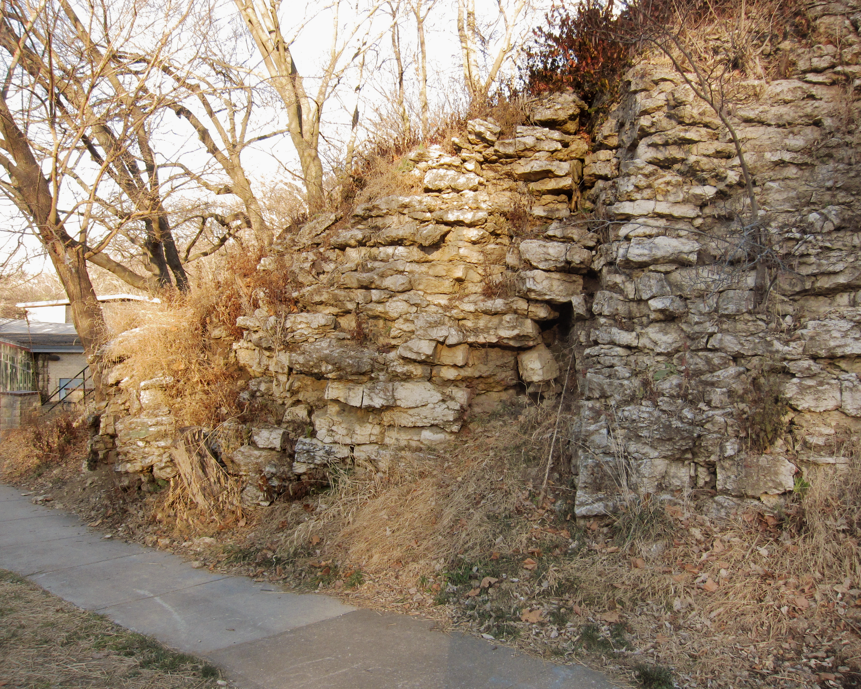 Stone ruins of stacked tan and grey limestone blocks form a tall wall alongside a concrete pathway, with bare winter trees and dried vegetation visible in the foreground and background.