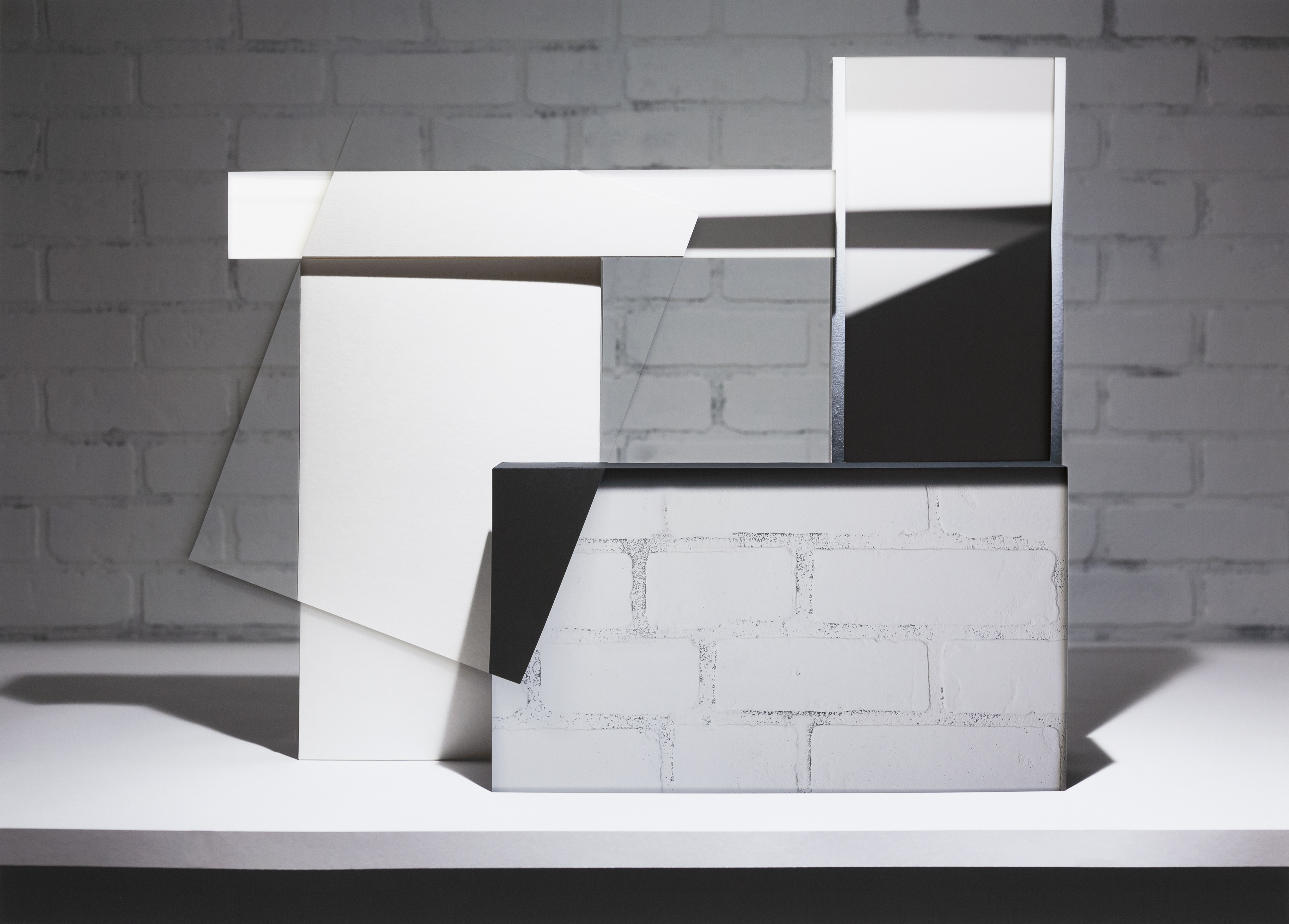 Abstract architectural composition featuring overlapping rectangular planes in white, black, and gray tones arranged in layered, three-dimensional forms against a white brick wall background. Strong diagonal lines and shadow play create depth across the stacked geometric elements.