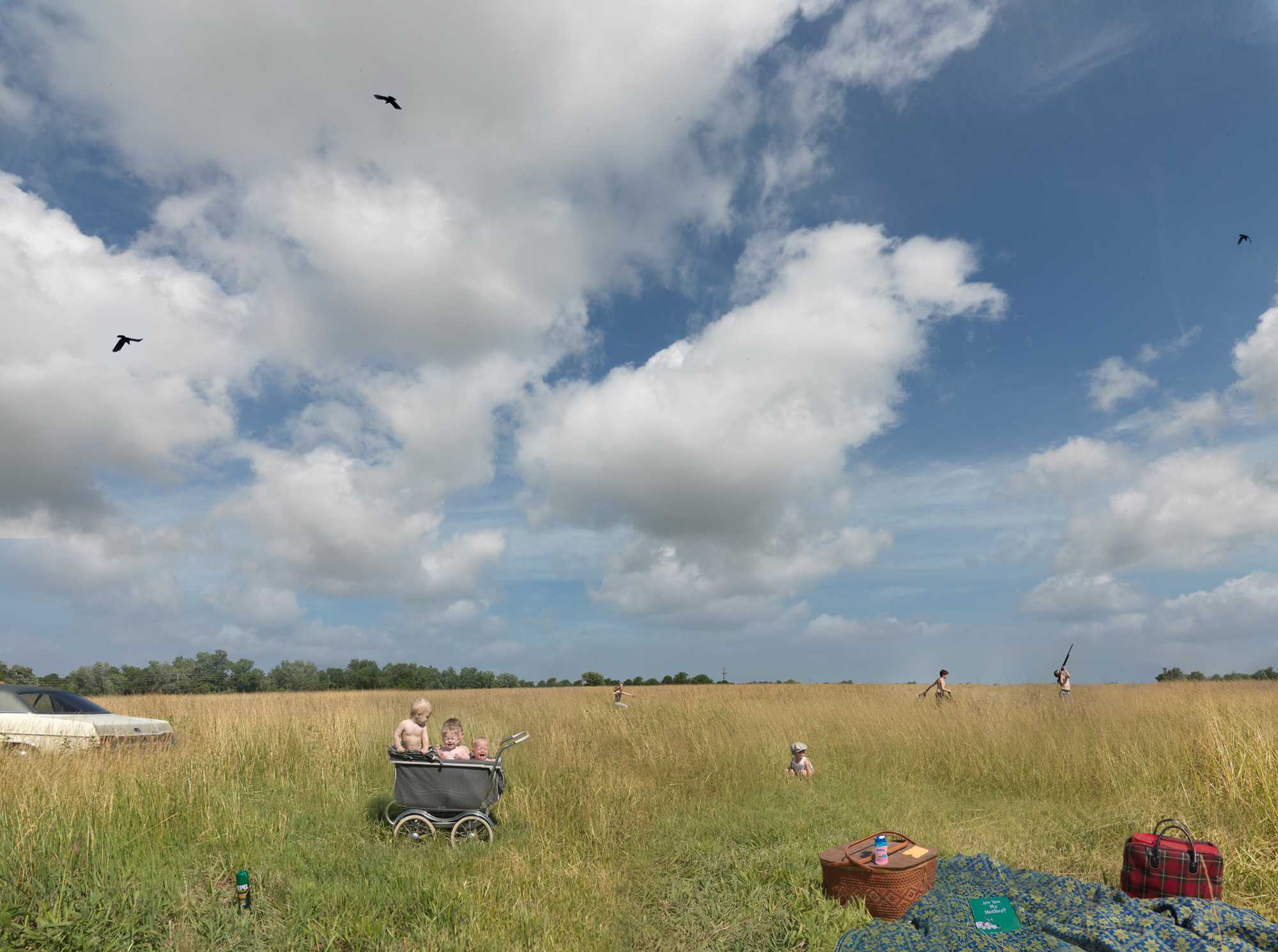 A expansive golden field under a partly cloudy blue sky, populated with figures engaged in leisure activities. In the foreground, a picnic setup with a wicker basket and plaid blanket sits on the grass, while a man pushes a child in a pram deeper into the field, accompanied by other figures flying kites and playing in the distance.