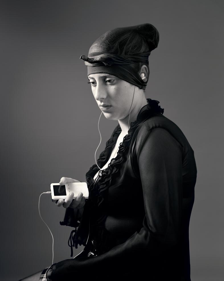 Black and white portrait of a woman in profile wearing a black hooded jacket and headband, holding a small white digital device with white earbuds. The minimalist composition is set against a neutral gray background with dramatic lighting that emphasizes the subject's contemplative expression.