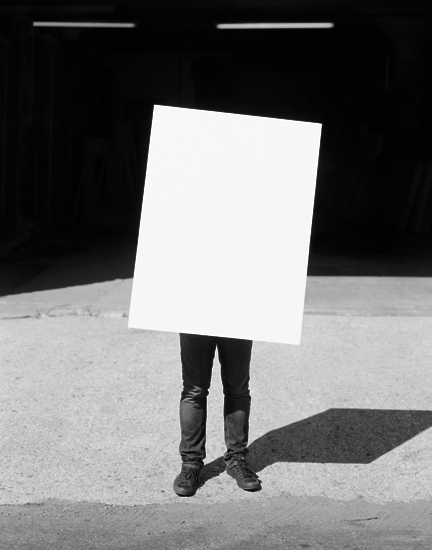 A figure in black clothing stands in an empty concrete courtyard holding a large blank white square panel in front of their head, obscuring their face. Their shadow is cast sharply on the ground beside them, while dark architectural walls frame the minimalist composition in black and white.