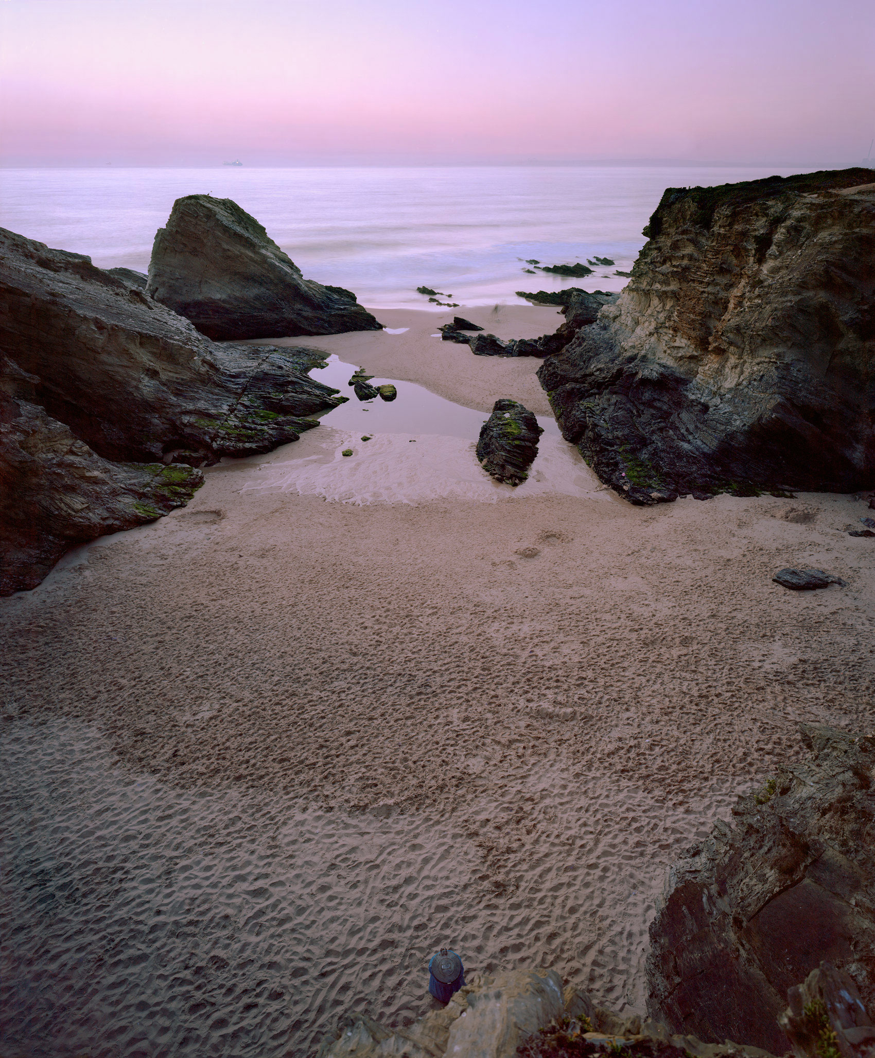 A coastal scene at dusk with dramatic rocky outcroppings framing a sandy beach and shallow pools of water. The sky glows in soft pink and lavender tones, while moss-covered stones and weathered rock formations create a sense of geological depth and layering across the composition.