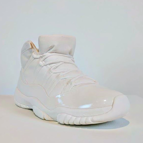 A white porcelain sneaker rendered in monochromatic tones, featuring a high-top silhouette with lacing detail and a glossy finish. The sculpture sits against a neutral background, casting a subtle shadow, with its smooth, ceramic surface capturing soft highlights.