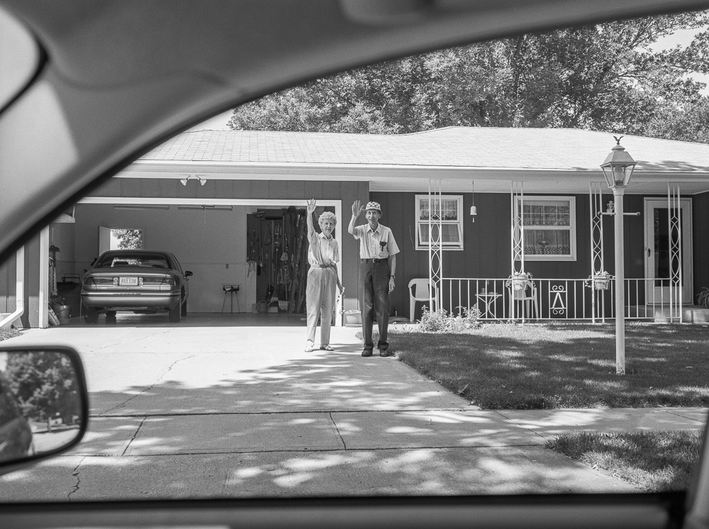 Black and white photograph taken from inside a car, showing a man and woman waving goodbye in the driveway of a single-story suburban house with an attached garage. The composition frames the scene through the car's windshield and side mirror, creating a layered perspective of the residential street and tree-lined background.