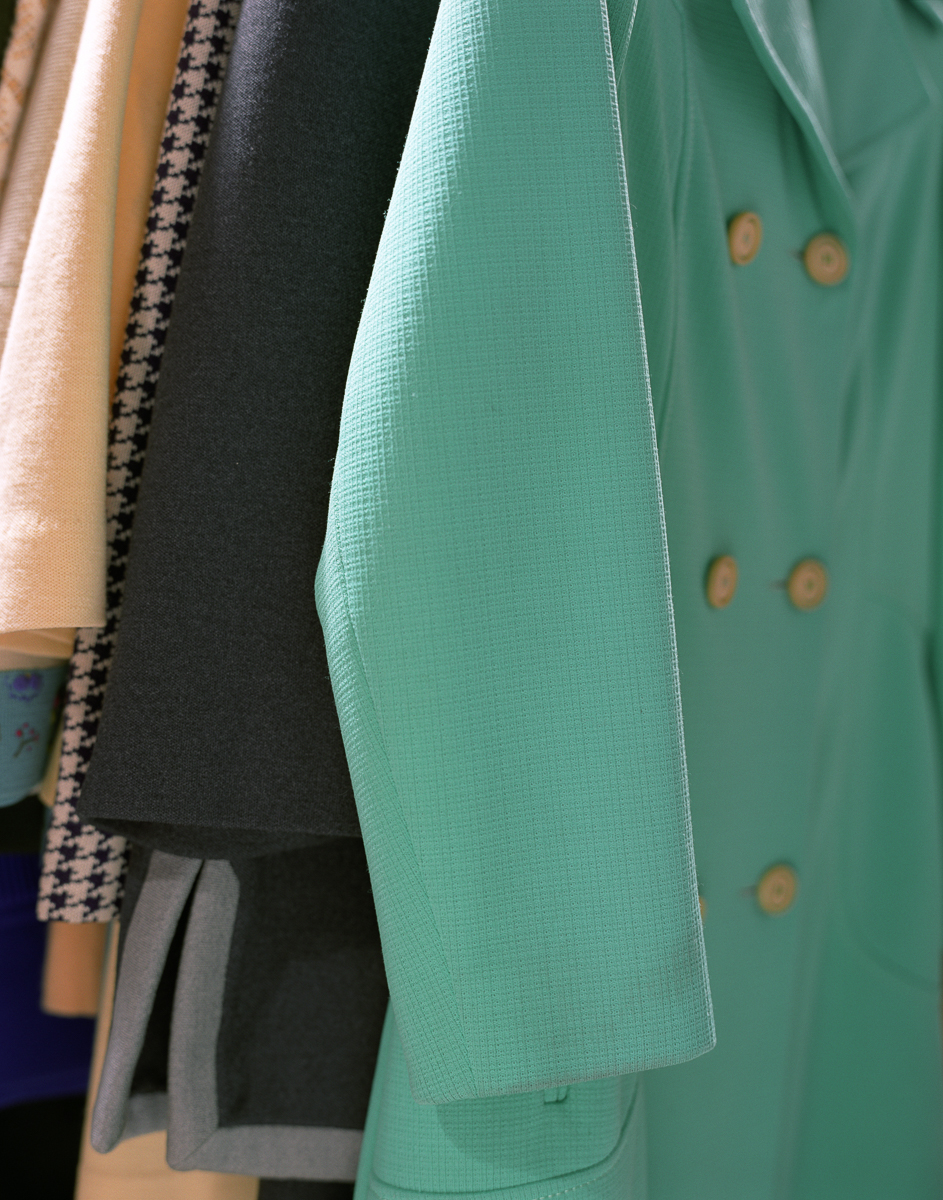 Close-up view of hanging garments displaying various textures and colors, including a mint green jacket with wooden button details, a dark charcoal piece, and lighter neutral tones, showcasing fabric weave and construction details.