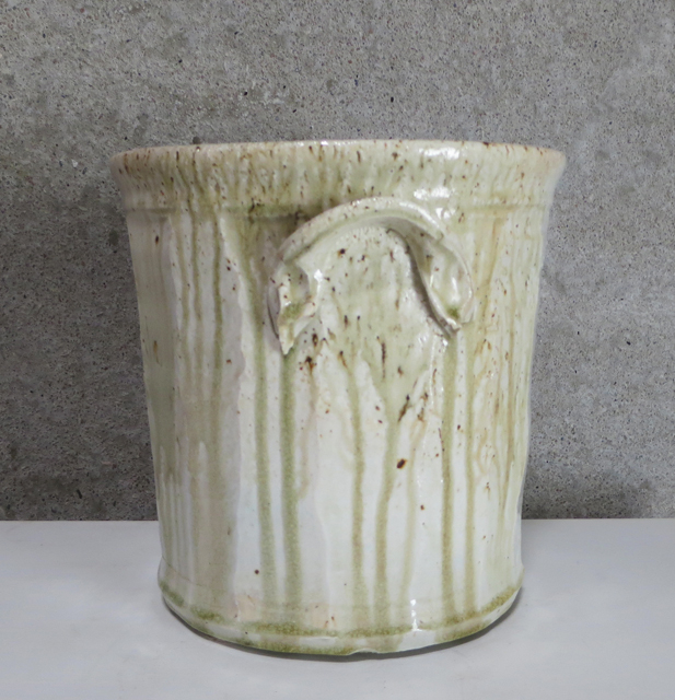 Cylindrical porcelain vessel with a cream-colored glaze featuring vertical striations and organic drip patterns, accented with brown speckles. The piece displays a subtle handle and exhibits the warm, natural variations characteristic of wood-fired ceramics.
