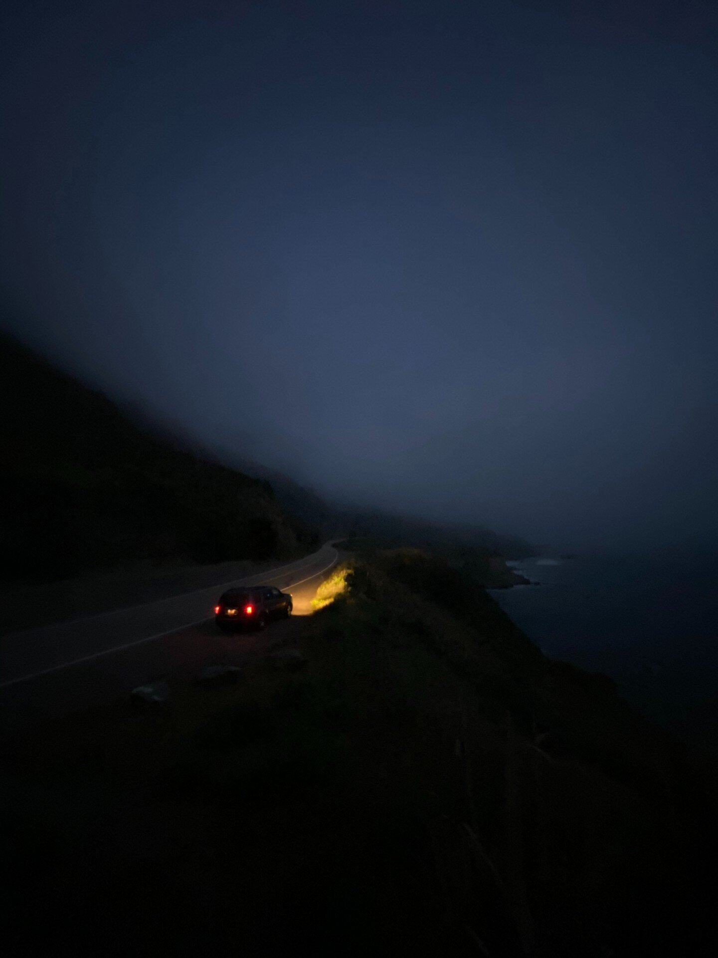 A vehicle with illuminated red taillights travels along a winding coastal road at dusk, its headlights casting a golden glow across the dark asphalt. Steep forested cliffs rise sharply on the left while the ocean and misty horizon occupy the right side, creating a moody, atmospheric scene rendered in deep blues and blacks with warm amber accents.