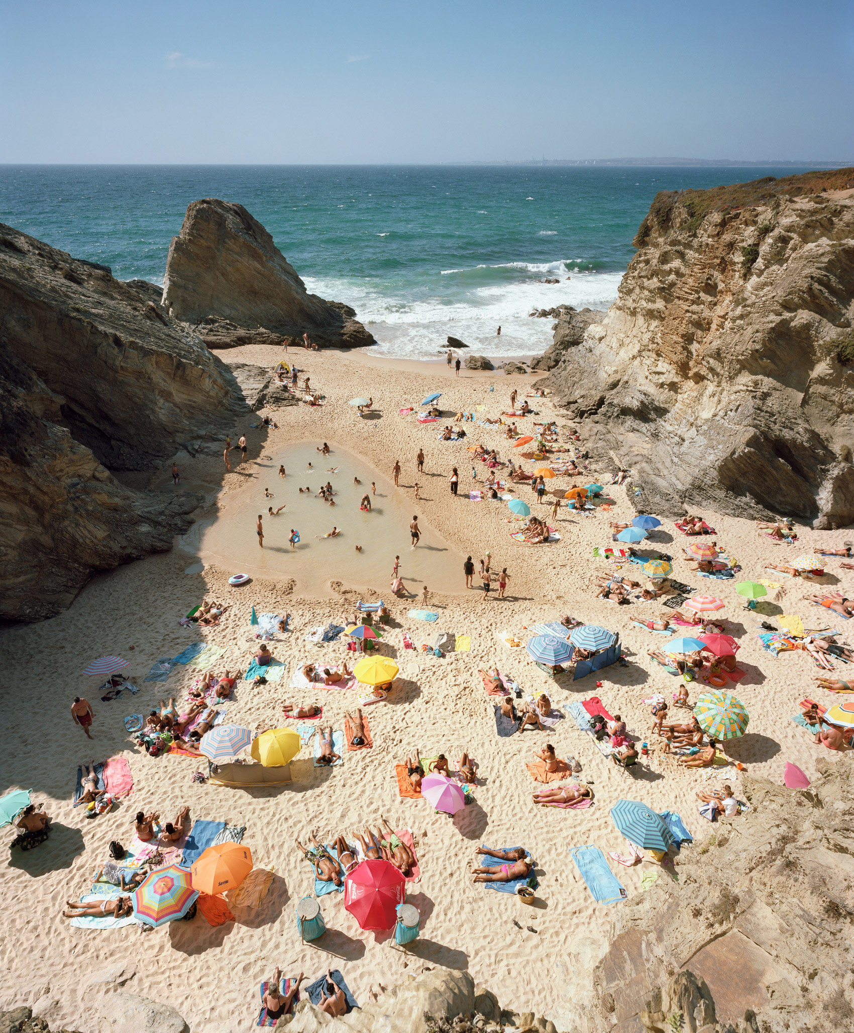 Aerial view of a crowded beach nestled between dramatic chalk cliffs, with numerous colorful umbrellas and sunbathers scattered across pale sand, turquoise waters, and breaking waves visible beyond.