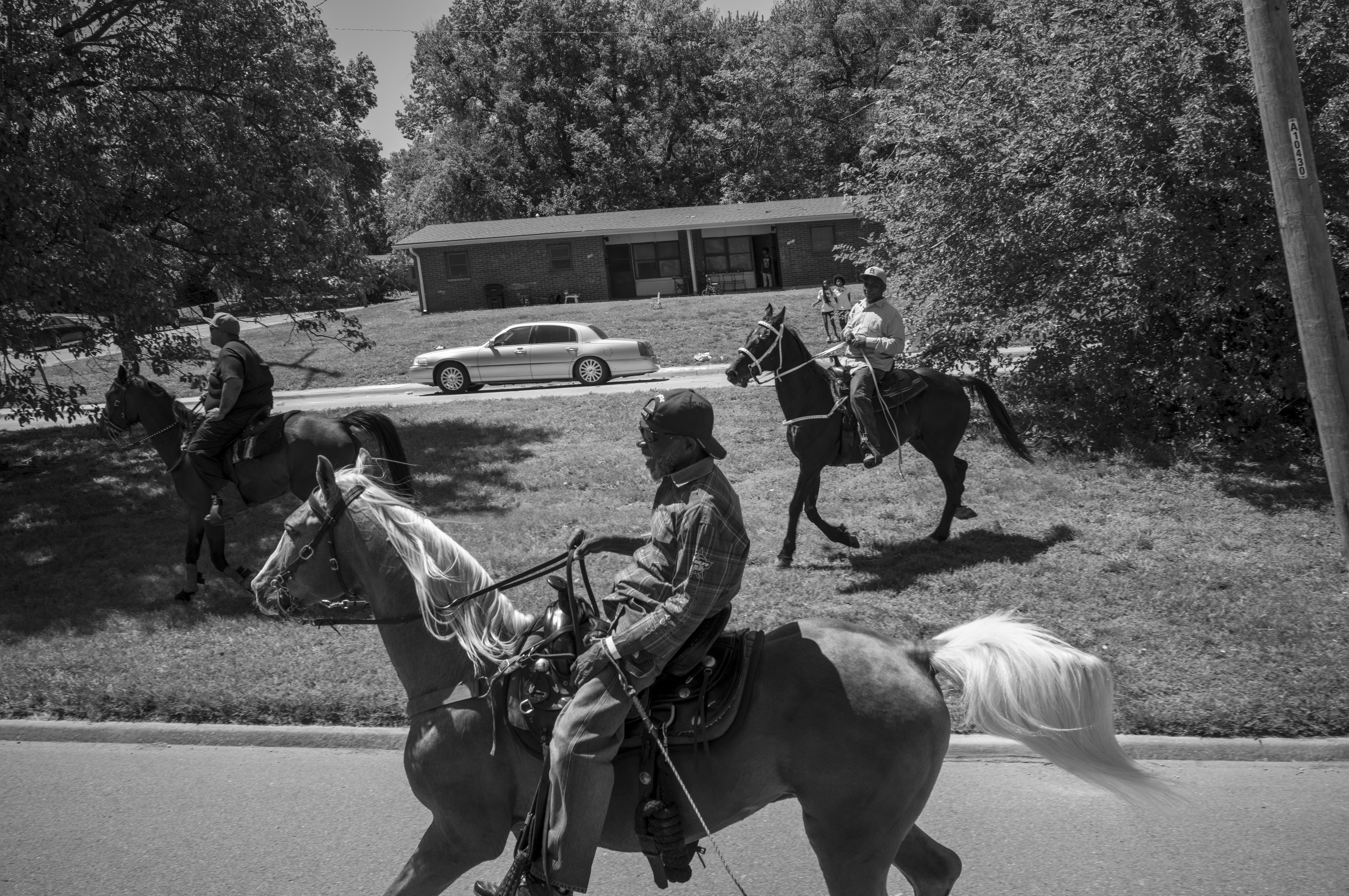 Black and white photograph depicting cowboys on horseback gathered in a residential driveway, with a silver sedan parked behind them and a modern brick house visible in the background, framed by dense foliage on either side.