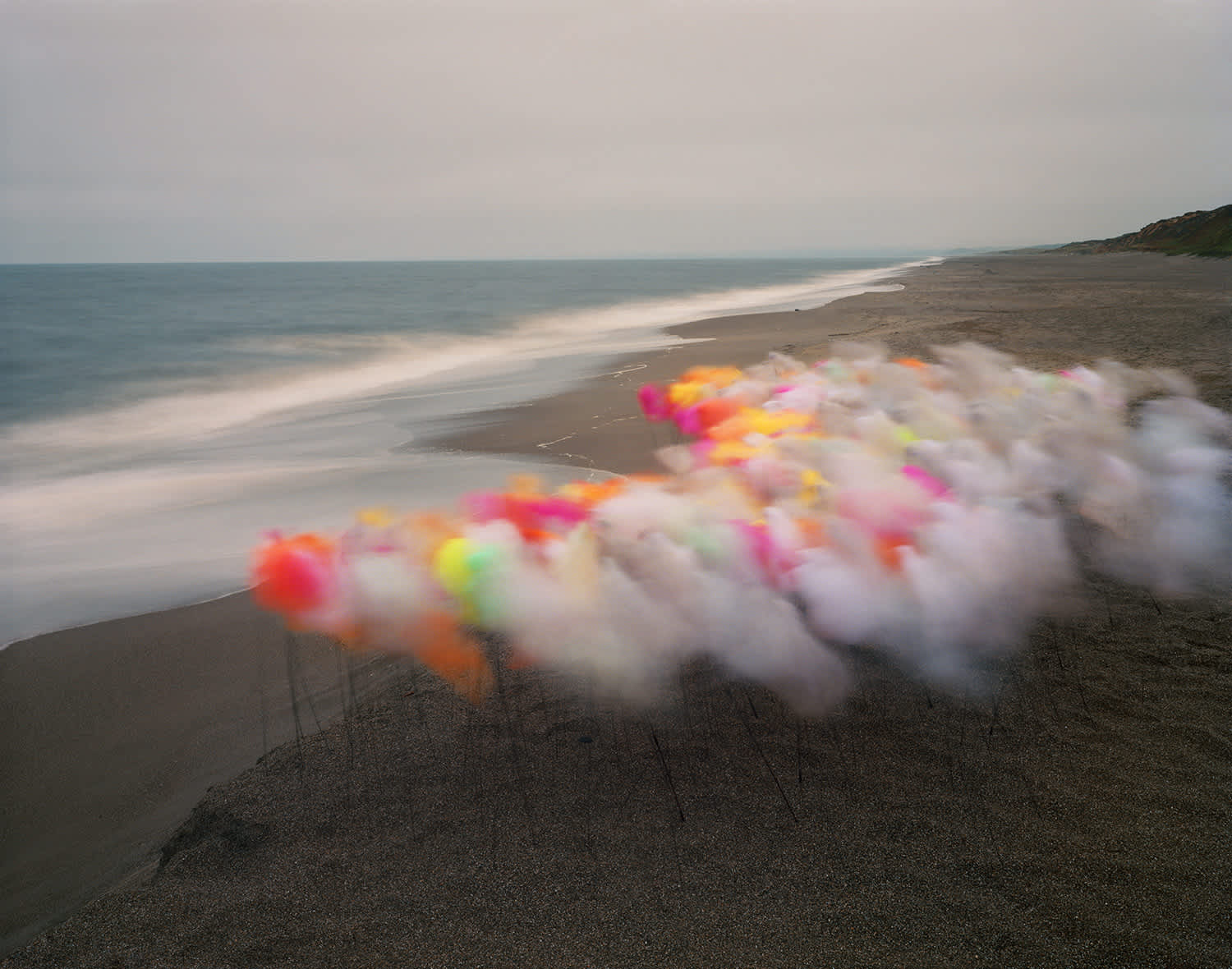 Blurred, colorful tulle fabric in shades of pink, yellow, orange, and red stretches across a dark sand beach, with a calm ocean and pale sandy shoreline visible in the soft-focus background.