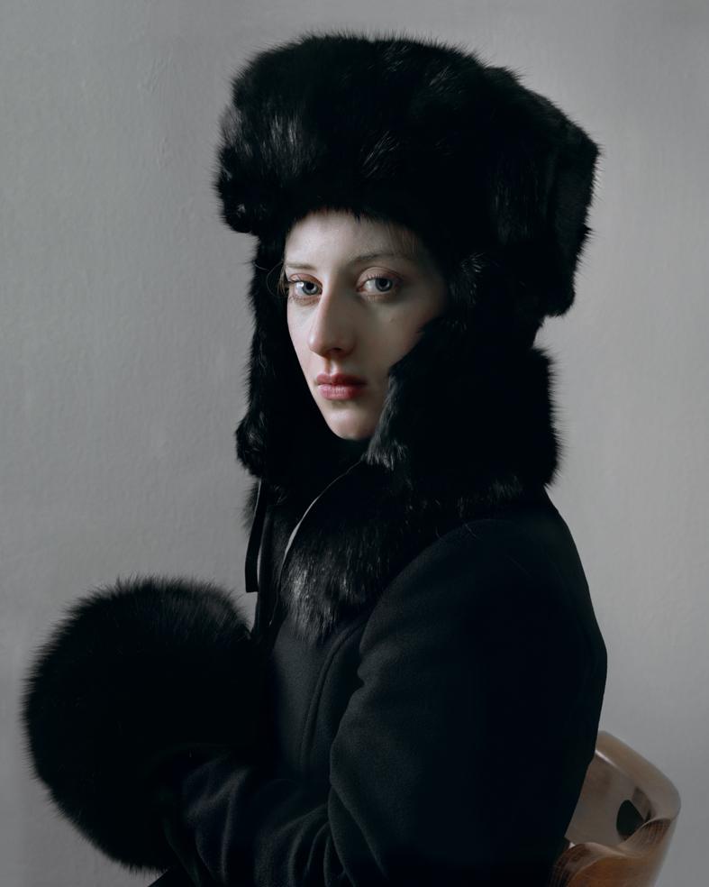 A person with pale complexion gazes over their shoulder while wearing a large black fur hat and collar against a neutral gray background. The subject is dressed in dark clothing with a striking fur muff held at their side, creating a composed portrait with dramatic contrast between the figure's fair skin and the rich black textures of the fur.