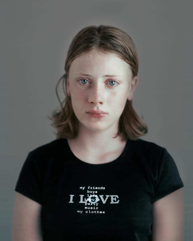 Portrait of a young person with shoulder-length brown hair wearing a black t-shirt printed with white text listing "my friends boys I LOVE party music my clothes" against a neutral gray background. The photograph employs a direct, frontal composition with a contemplative expression and cool color palette.