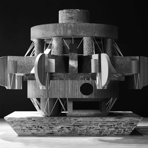 Black and white photograph of a geometric sculptural structure with cylindrical and angular forms, featuring stacked metal elements, curved discs, and triangular bracing against a dark background. The composition is symmetrical and frontal, showcasing industrial materials and architectural precision with strong contrasts of light and shadow.
