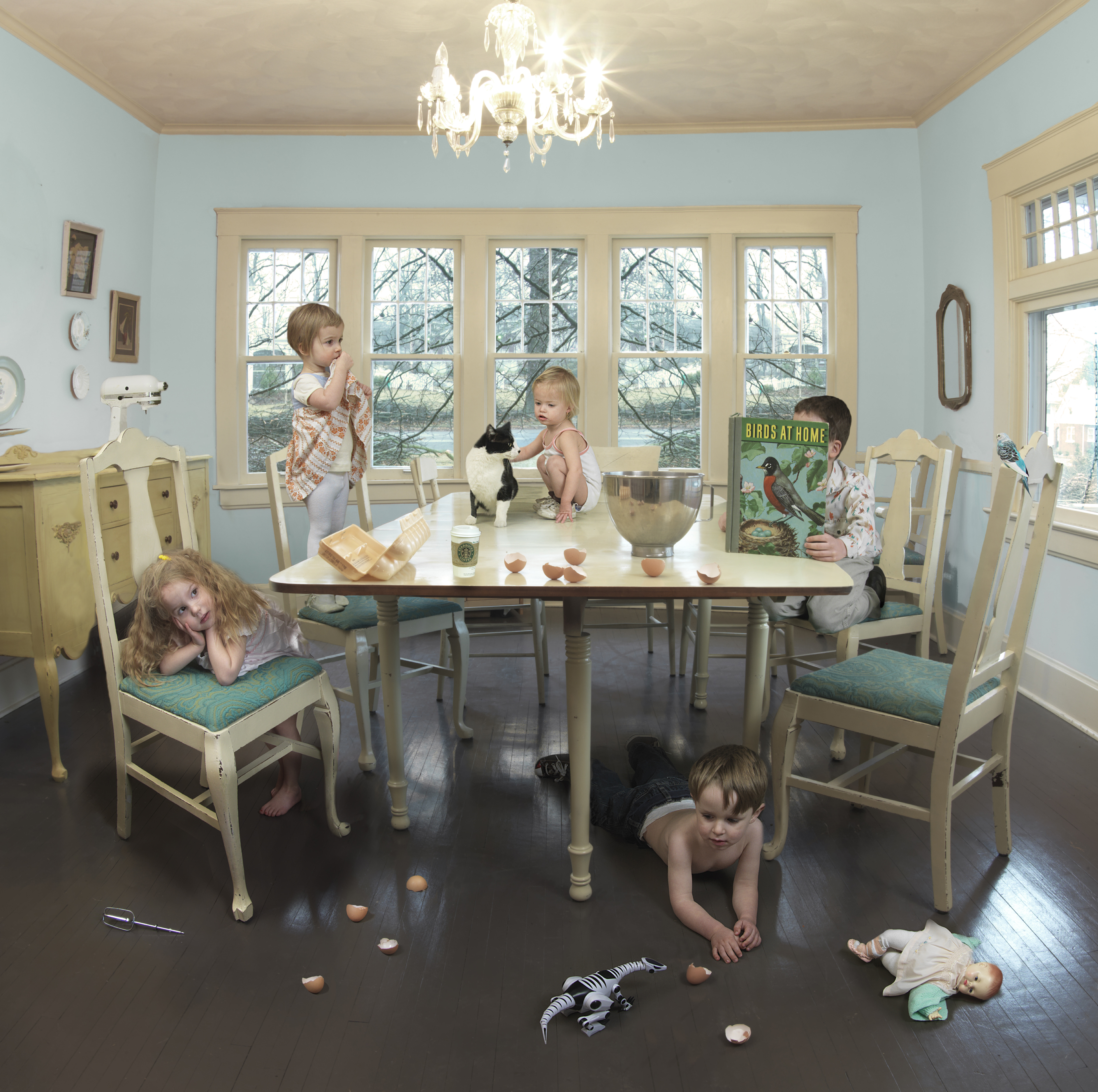 A staged domestic scene shows multiple young children and black cats engaged in playful chaos around a cream-colored dining table in a light blue room with large windows. Children are seated at chairs, crawling on a dark wooden floor scattered with nuts, and interacting with the cats, while a metal bowl and other objects sit on the table, creating a carefully composed tableau of controlled disorder.
