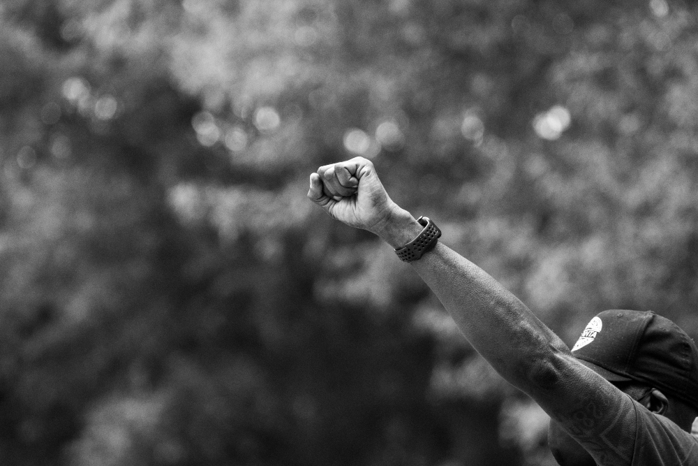Black and white photograph of a raised fist against a blurred natural background. The arm extends diagonally across the frame, wearing a dark sleeve and a beaded bracelet, captured in sharp focus while the foliage behind remains soft and out of focus.