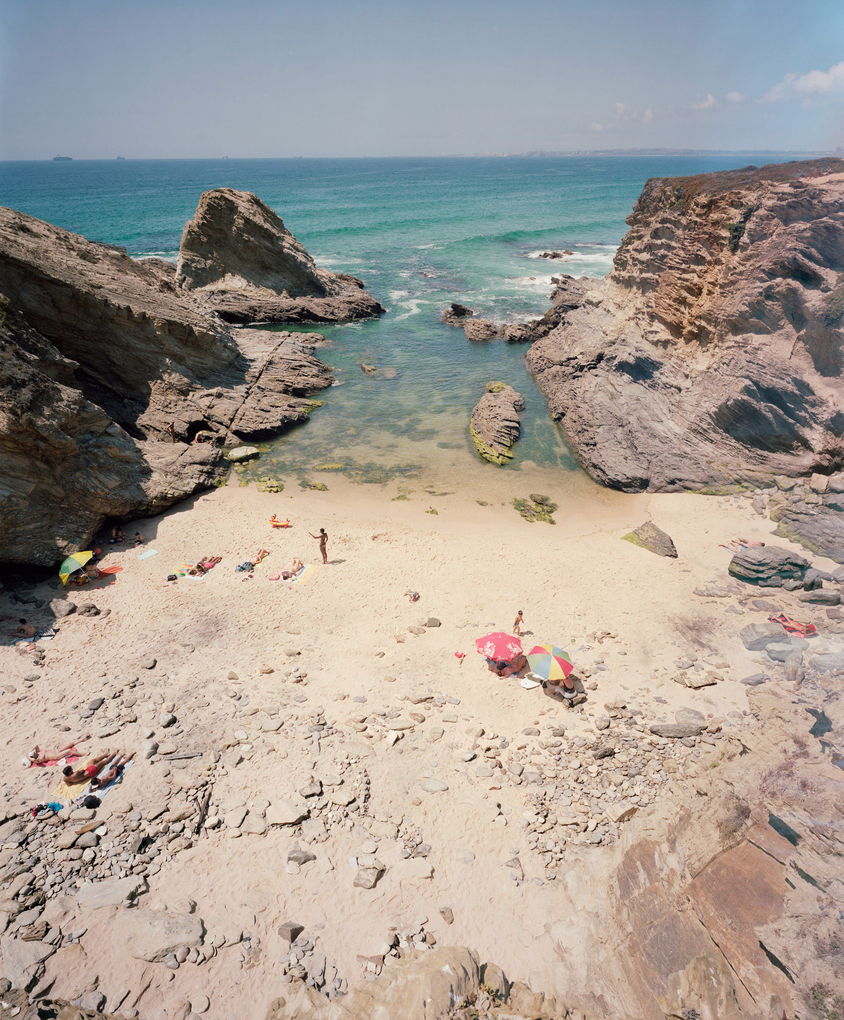 A secluded cove with turquoise waters framed by dramatic rocky outcrops, featuring a sandy beach dotted with colorful beach umbrellas and sunbathers scattered across the shore. The composition captures the scene from an elevated vantage point, emphasizing the contrast between the pale sand, crystalline water, and weathered stone formations under bright daylight.