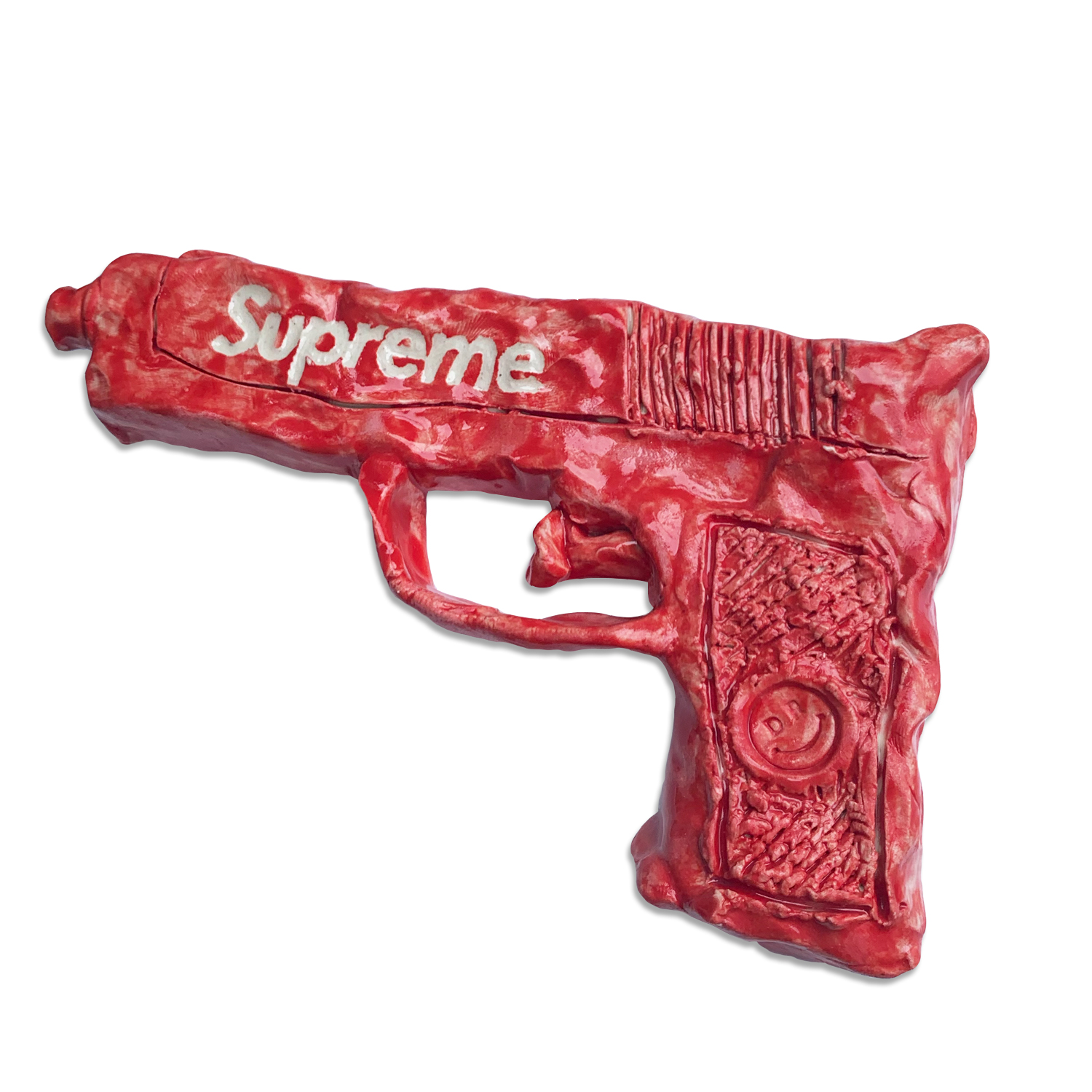 A bright red ceramic sculpture of a toy pistol, featuring the word "Supreme" embossed across the barrel. The piece displays intricate surface detailing and texturing on the grip, rendered in glossy glazed clay.