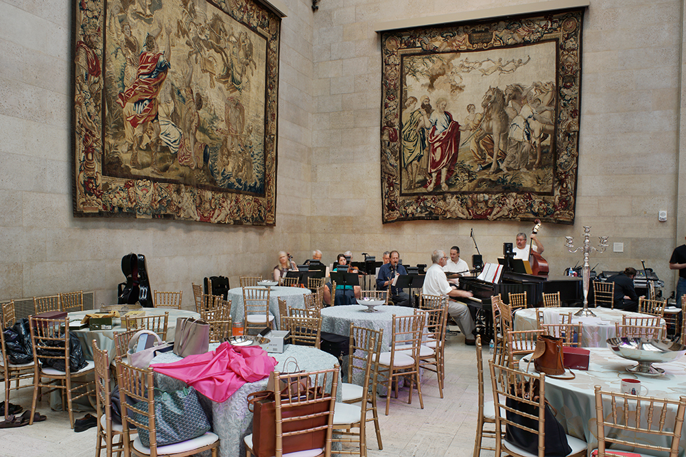 A grand hall with ornate tapestries depicting classical scenes hangs above round dining tables set with white linens, wooden chairs, and colorful fabric accents. A grand piano and musicians are visible in the background as event preparations take place.