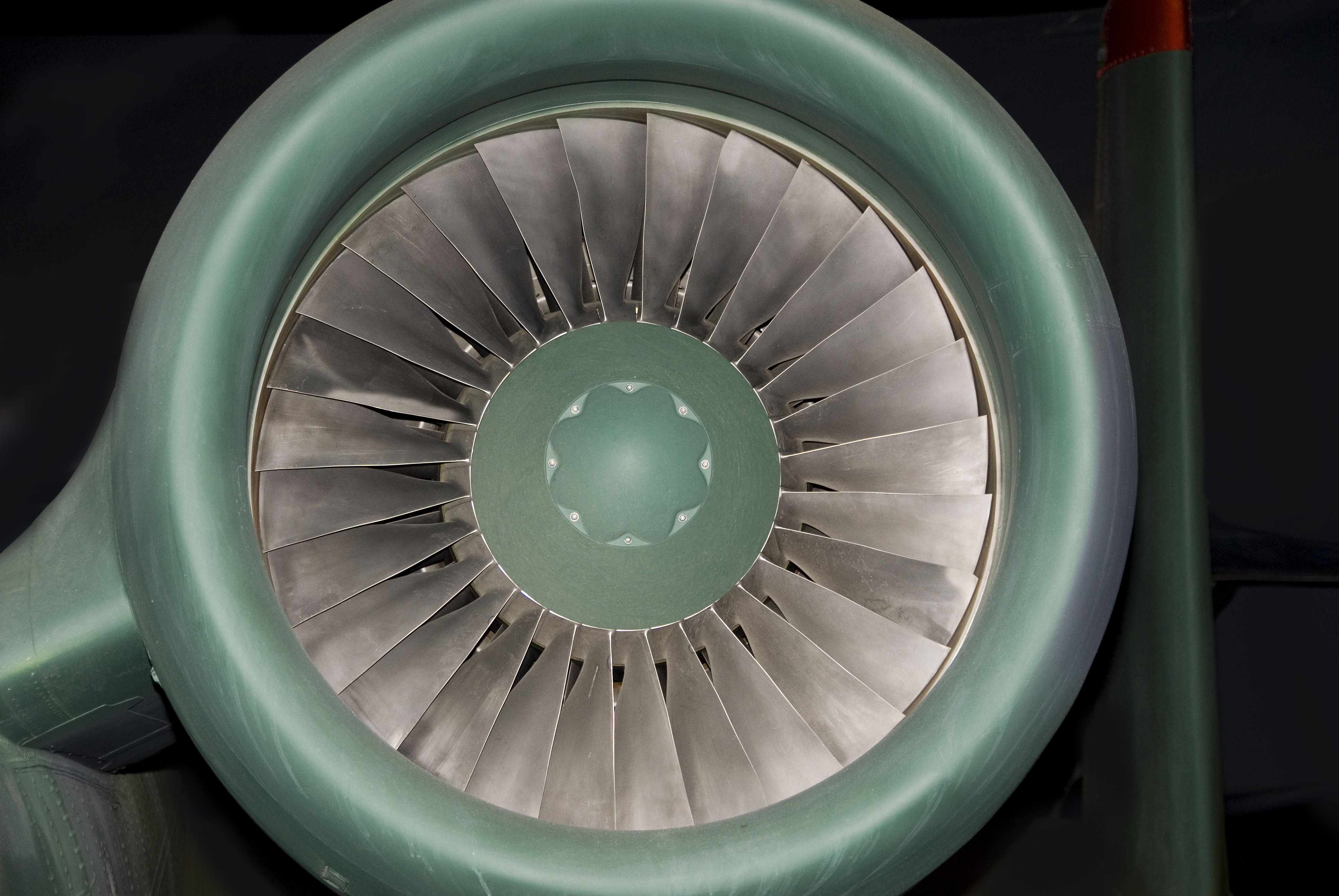 Frontal view of a jet engine intake with concentric metal fan blades radiating from a central green hub, set against a dark background. The polished steel blades create a symmetrical, geometric composition in silvery tones contrasted with the muted sage green of the engine casing.