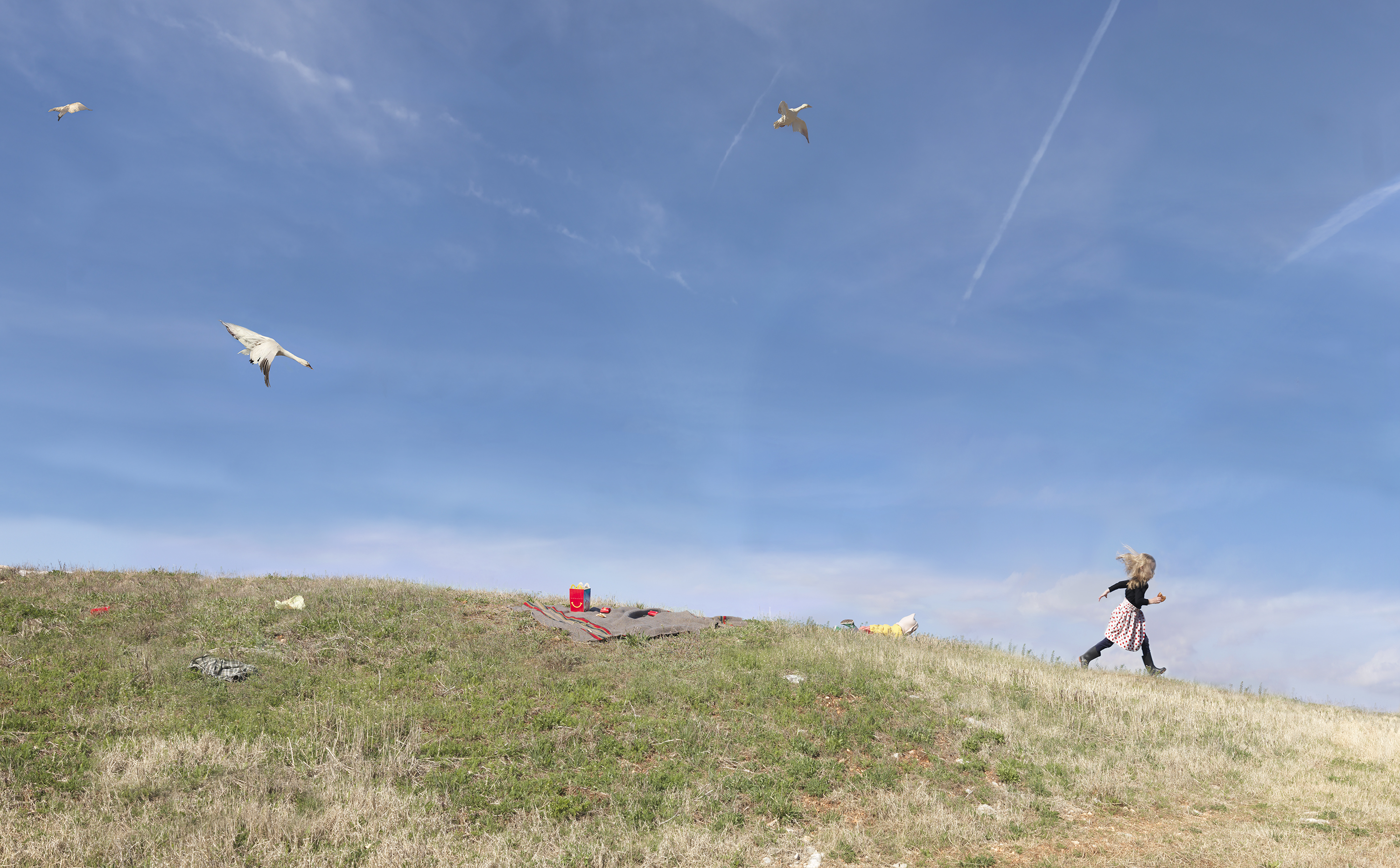 A person in a patterned dress runs across a grassy hillside while multiple kites fly overhead in a bright blue sky, with scattered picnic items visible on the ground below.
