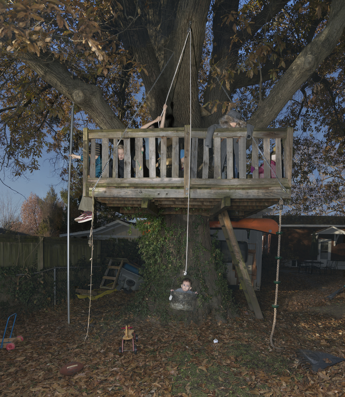 A wooden treehouse built high in a large tree branches over a backyard scene. Children visible both inside the railed platform and swinging from ropes below, surrounded by autumn foliage, fallen leaves, and residential structures in soft afternoon light.
