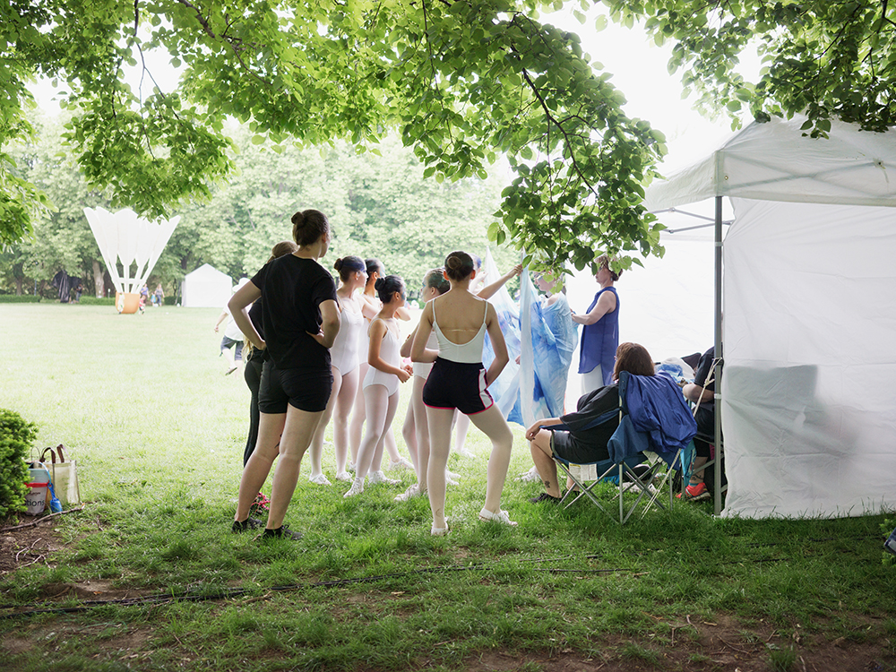 A group of young dancers in ballet attire gather beneath leafy trees in a grassy park setting, with a white tent and spectators visible in the background. The scene captures a moment of community and performance in an outdoor natural environment.