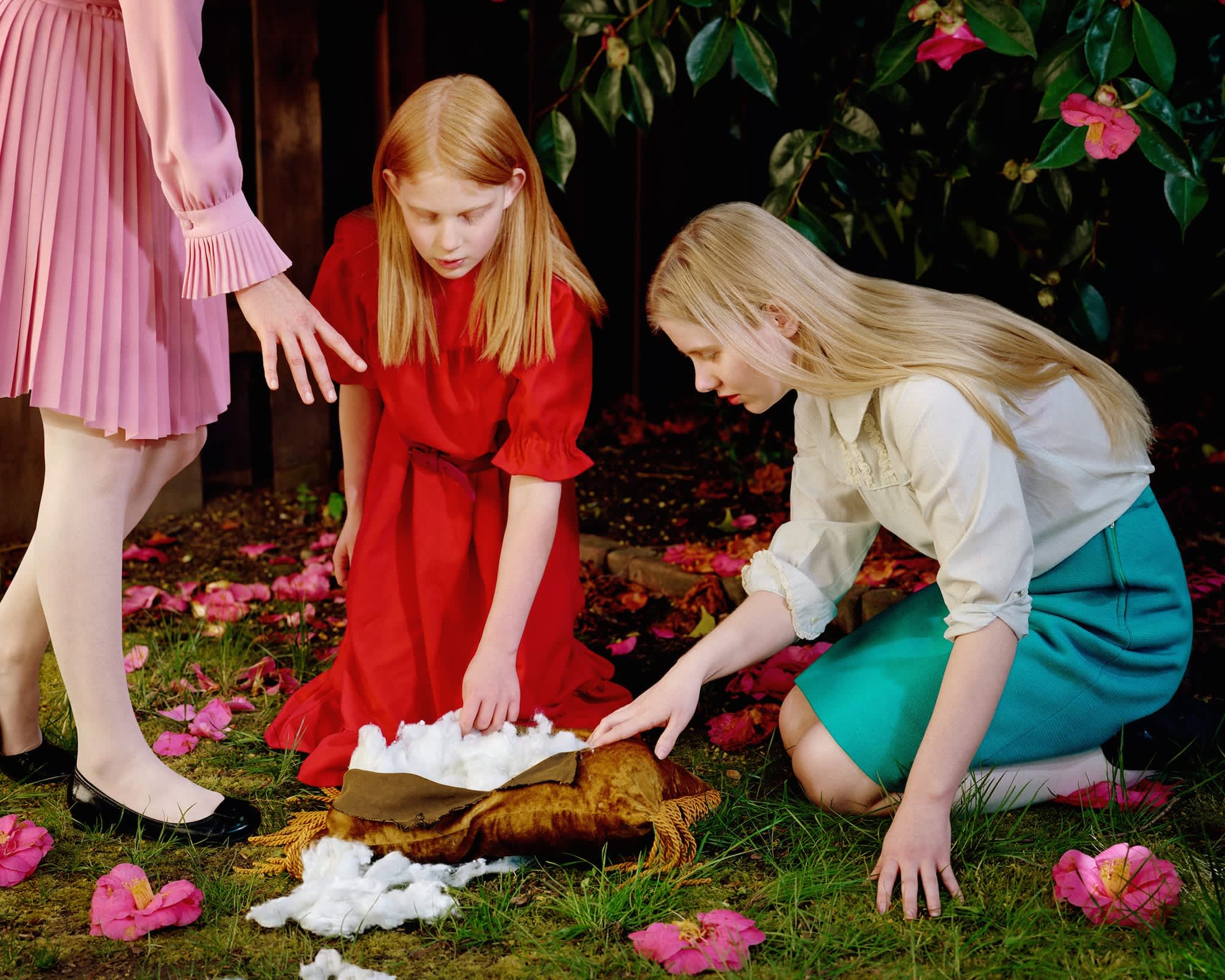 Three young girls in a garden setting examine a golden-brown sculptural form resting on white fabric scattered across moss and grass. Pink flowers bloom throughout the scene, while an adult in a pink pleated skirt stands nearby, and climbing vines with pink flowers frame the composition against a dark background.
