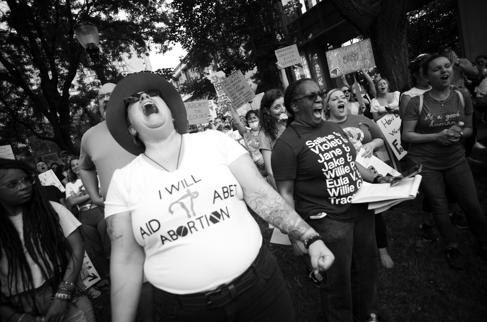Black and white photograph of a pro-choice protest in an urban park setting. A woman in the foreground wears a white t-shirt with text reading "I WILL AID ABET ABORTION" and a wide-brimmed hat, her head tilted back in an expressive shout. She is surrounded by other protesters holding signs, some visible in the background among trees, captured in a moment of passionate activism and collective demonstration.