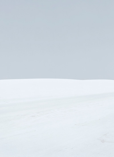 Minimalist landscape divided by a subtle curved horizon line, with a pale blue-grey sky occupying the upper portion and a soft white snow-covered ground below, rendered in muted tones with delicate tonal variations.