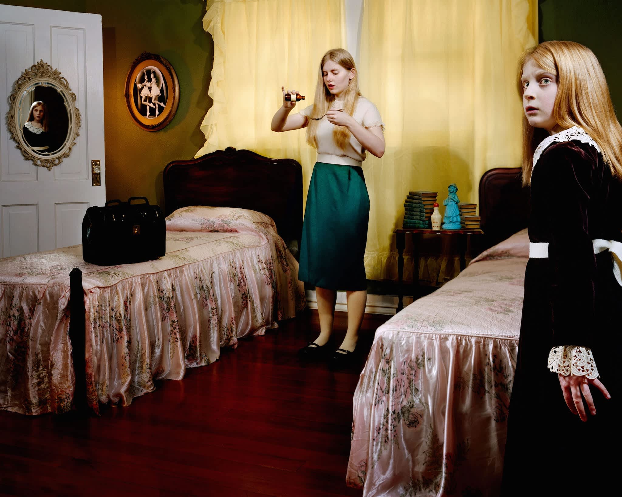 Two young girls with reddish-blonde hair in a yellow-walled bedroom with twin beds covered in floral patterned spreads. One girl in the foreground wears a dark velvet dress with lace cuffs, gazing toward the camera with a contemplative expression. The other girl stands in the center holding a small dark object, wearing a green skirt and light top. The room contains vintage furnishings including ornate mirrors, a dark wooden dresser with stacked books and blue decorative objects, a suitcase, and wall-mounted artwork. The composition employs warm, saturated colors and precise, staged composition characteristic of contemporary color photography.