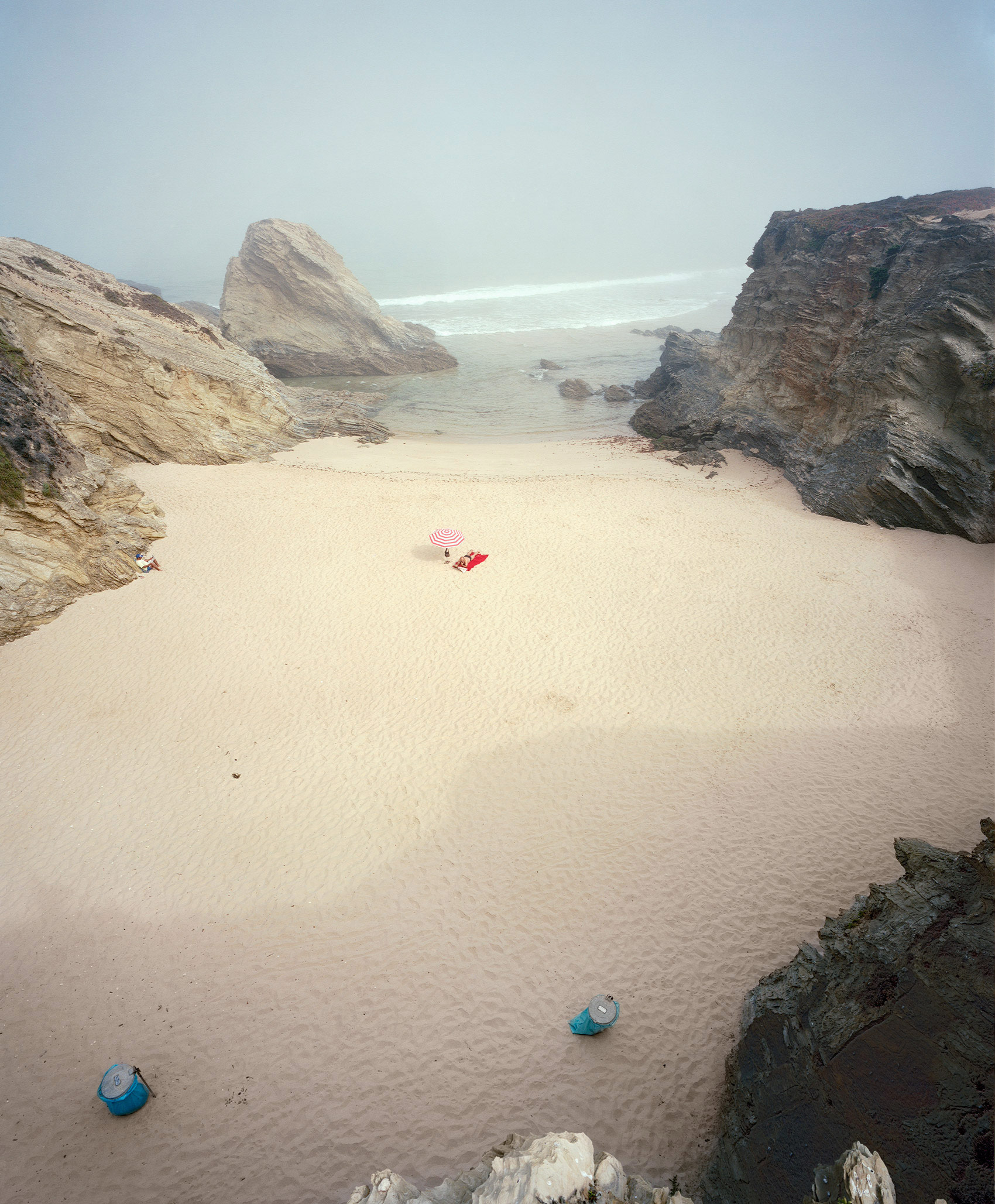 A secluded sandy beach enclosed by dramatic rocky outcrops, viewed from an elevated vantage point. A few colorful beach umbrellas and towels dot the pale sand near the water's edge, while the turquoise sea stretches toward a hazy horizon under an overcast sky.