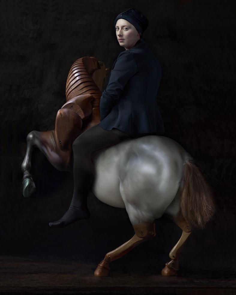 A woman in dark clothing and a dark headwrap sits atop a rearing wooden rocking horse against a black background. The horse, rendered in rich brown tones with a white blaze on its chest, is captured mid-motion, while the woman gazes toward the viewer with a composed expression.