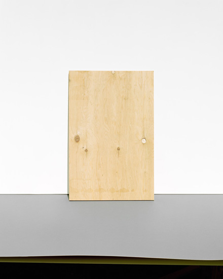 A light wooden rectangular panel leans against a gray wall above a dark horizontal stripe, positioned in a minimalist composition with a plain beige background.