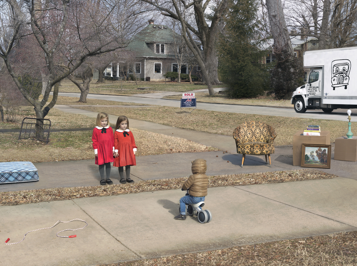 A residential driveway scene with a young child riding a tricycle in the foreground, wearing a tan jacket and blue pants. Two girls in matching red dresses stand in the middle distance watching. A "SOLD" real estate sign, furniture, books, and household items are arranged on the driveway as if for a garage sale or moving day. A moving truck and bare trees frame the suburban house behind them.