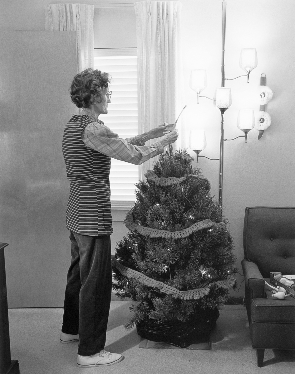 Black and white photograph of a woman in a striped shirt standing beside an unconventional Christmas tree made entirely of evergreen branches and rope, decorated with modern glass pendant lamps that hang from above. She holds a newspaper and gazes at the sculptural tree installation in a minimalist interior space with white curtains and mid-century modern furniture.