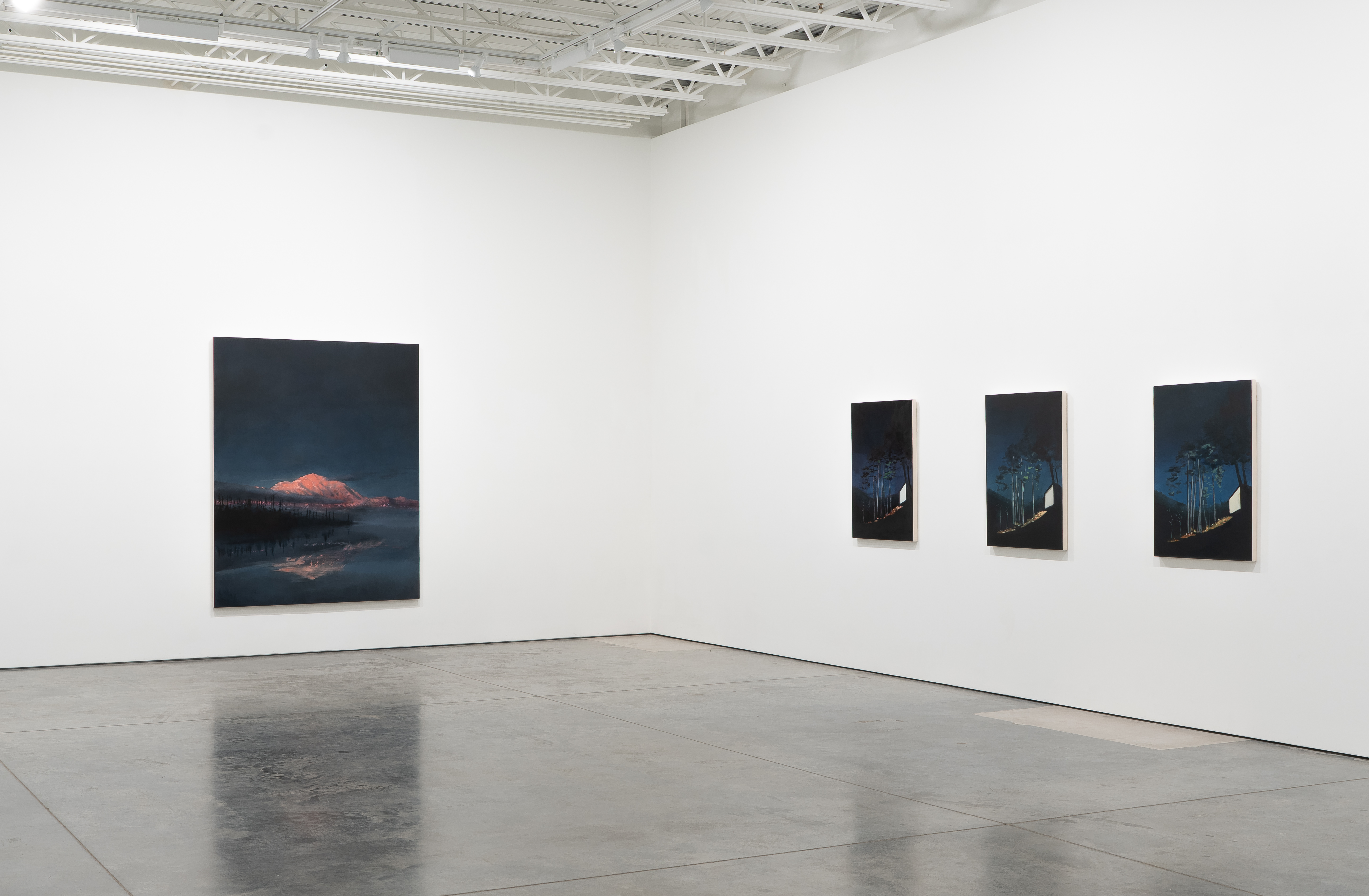 Gallery interior with four paintings hung on white walls under industrial ceiling. On the left, a large square canvas depicts a snow-capped mountain glowing red at dusk, reflected in still water beneath a dark blue sky. On the right wall, three smaller vertical canvases show nocturnal forest scenes with pale geometric shapes suggesting structures or clearings against dense dark trees.