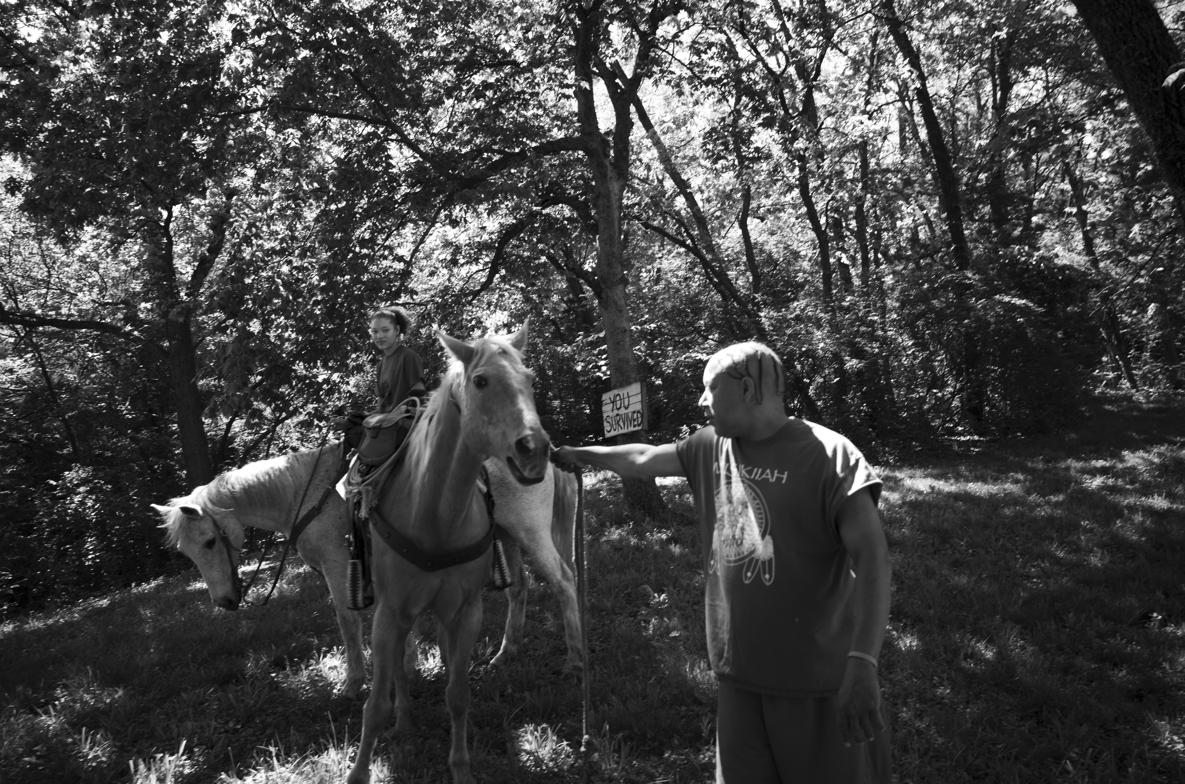 Black and white photograph of two figures in a wooded forest setting with a saddled horse between them. The figure on the right wears a graphic t-shirt and reaches toward the horse, while another figure is visible mounted or positioned on the horse in the background. Dense trees and dappled light fill the composition, creating a naturalistic woodland scene.