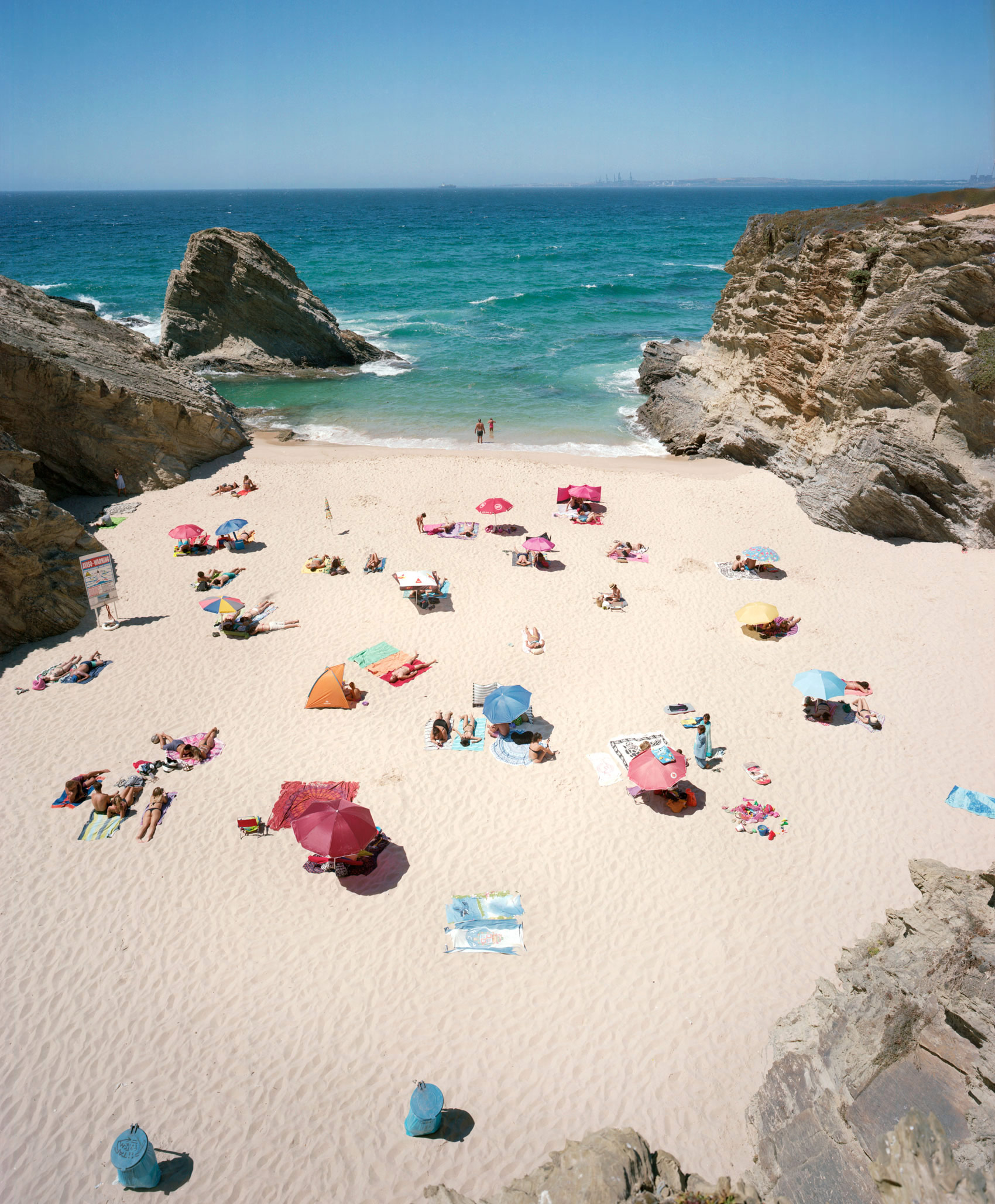 Aerial view of a sandy cove beach flanked by dramatic rocky outcrops, with numerous beachgoers scattered across the sand under colorful umbrellas and beach tents in shades of pink, blue, yellow, and orange. Turquoise water with white-capped waves frames the scene, with a few swimmers visible near the shore.
