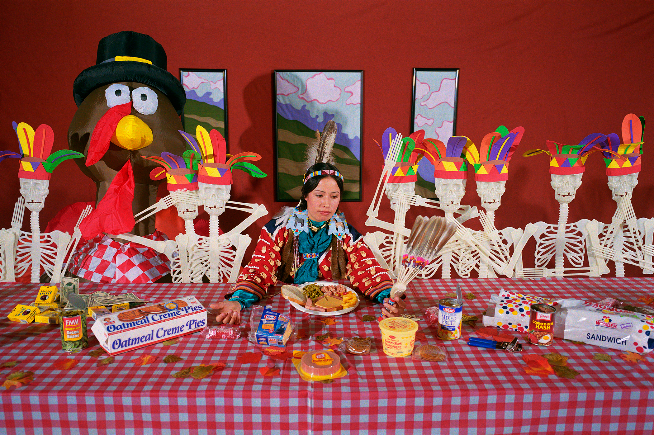 A woman in traditional Native American regalia sits at a red and white checkered table laden with processed foods and snack items, flanked by skeleton figures with colorful paper flower headdresses against a vibrant red wall. White skeletal remains frame the scene alongside a large caricatured bird figure wearing a top hat, creating a satirical juxtaposition of indigenous imagery, consumer culture, and memento mori symbolism.