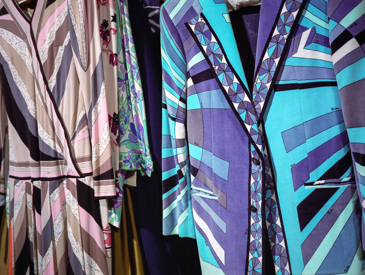 Colorful printed garments displayed on hangers, featuring bold geometric patterns and vibrant hues of turquoise, purple, pink, and cream. The designs incorporate repeating motifs including angular stripes, floral elements, and ornamental borders characteristic of mid-century fashion design.