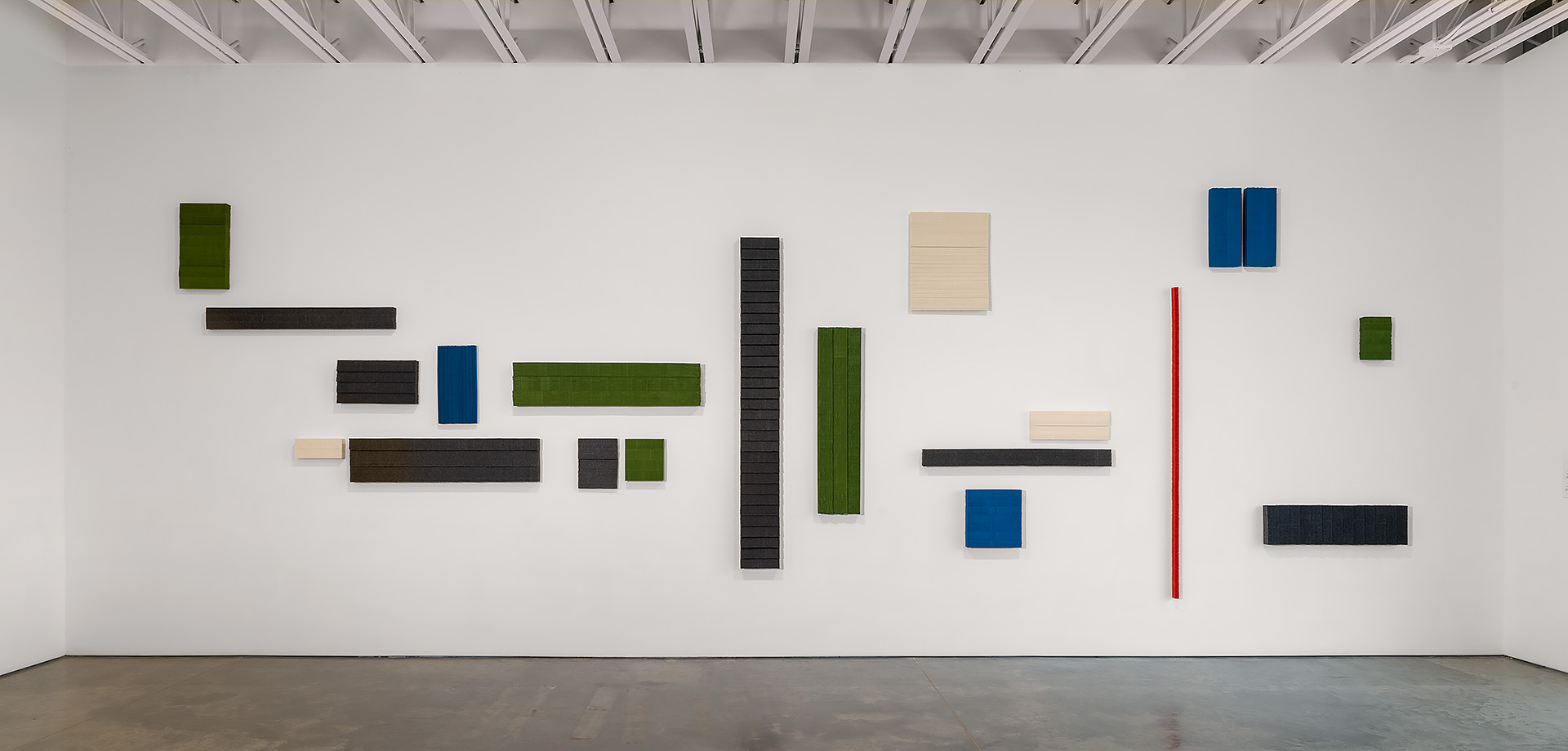 Abstract wall installation composed of rectangular and square panels in varying sizes arranged across a white gallery wall. Panels in dark charcoal, forest green, cream, blue, and a single red vertical element create a rhythmic composition against the minimalist white backdrop.