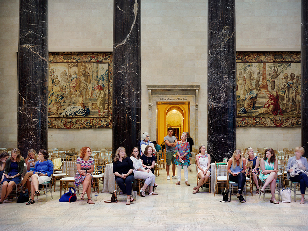 A group of seated women arranged in a semicircle on a polished floor within a grand hall featuring towering black marble columns, classical tapestries depicting figures in period dress, a golden arched doorway, and cream-colored walls.