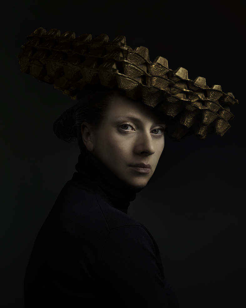 Portrait of a woman in profile against a dark background, wearing an elaborate sculptural headdress composed of layered gold-leaf geometric forms. She is dressed in black, her gaze directed downward and away, lit by subtle side lighting that emphasizes the dimensional quality of the metallic headpiece and creates a contemplative mood.