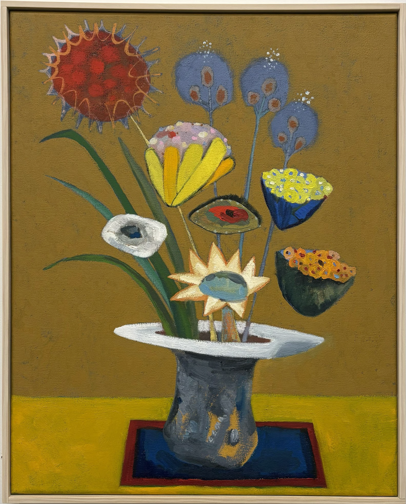 A surreal still life painting featuring a dark ceramic vase with a white-rimmed opening, filled with an imaginative arrangement of stylized flowers and botanical forms. The composition includes a red spiky flower, white anemone, yellow petals, blue clustered blooms, and small potted plants with yellow and orange flowers emerging from the vase, set against a golden-brown background with a yellow-green floor and red-blue base.