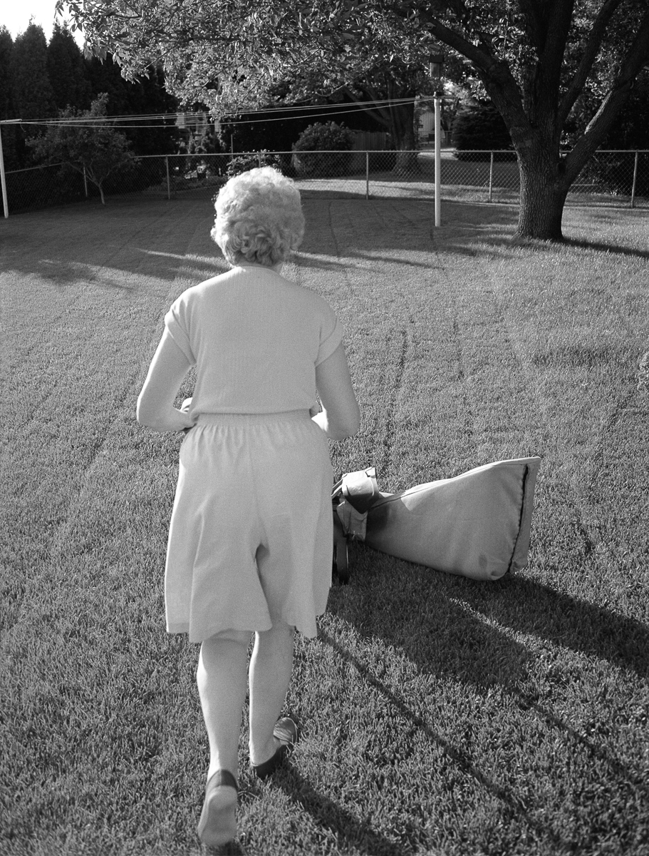 Black and white photograph of a person with white hair jogging across a grassy yard while carrying a rolled-up lawn mower, with a tennis court and trees visible in the background.